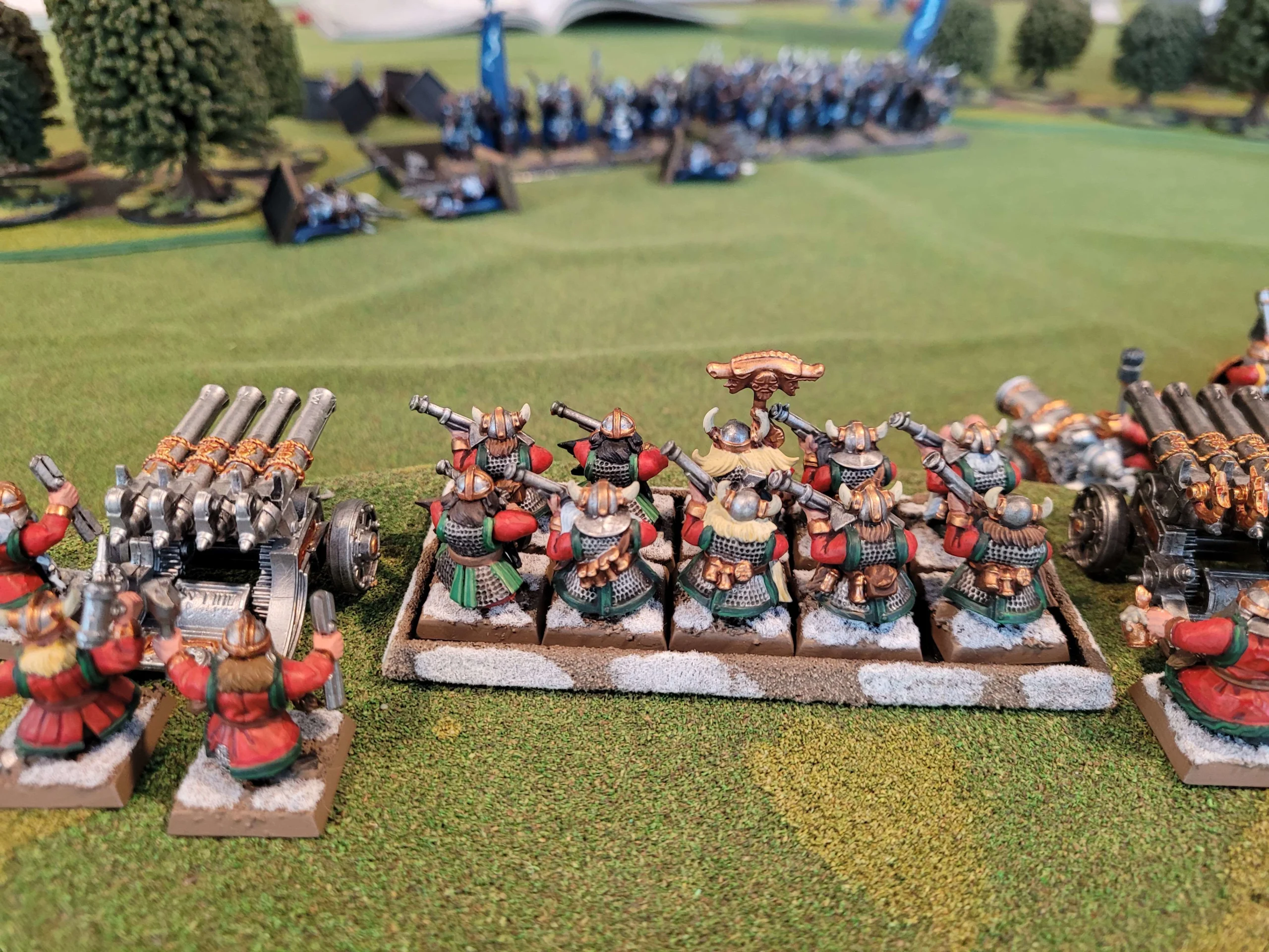 Close up photo of a hill on a Warhammer table that's occupied by a unit of ten dwarf thunderers, and two organ guns. They're overlooking a valley occupied by Tzeentchian Chaos Warriors who are suffering casualties from gunfire.